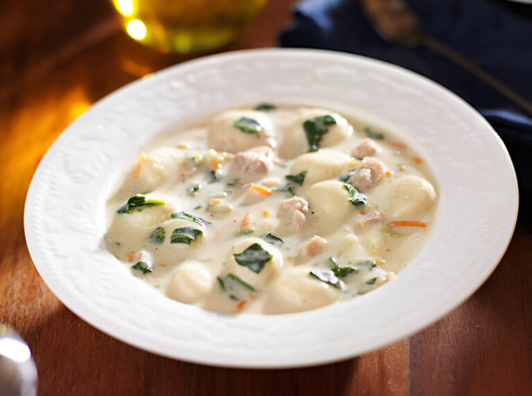 A creamy soup with gnocchi, vegetables, and pieces of meat served in a decorative white bowl.