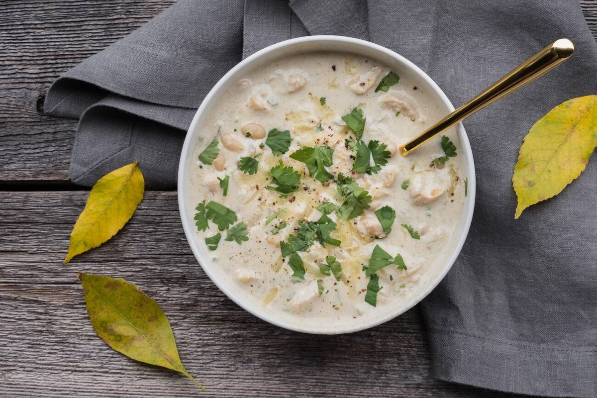 A bowl of creamy soup garnished with fresh herbs, placed on a wooden surface with a grey napkin and autumn leaves.