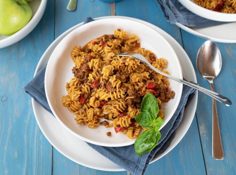 A plate of spiral pasta with ground meat and diced red peppers, garnished with fresh basil.