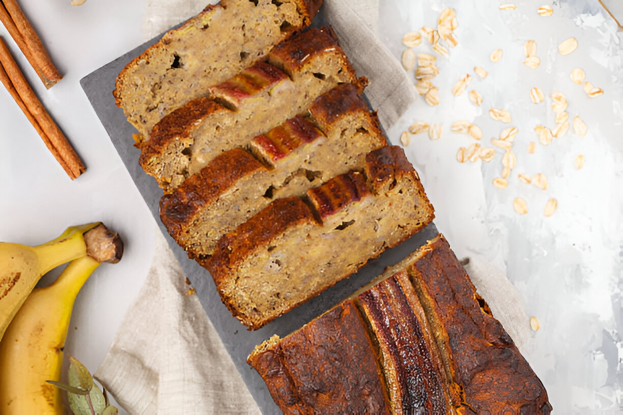 A freshly baked banana bread loaf sliced on a dark slate board, with whole bananas and cinnamon sticks nearby.