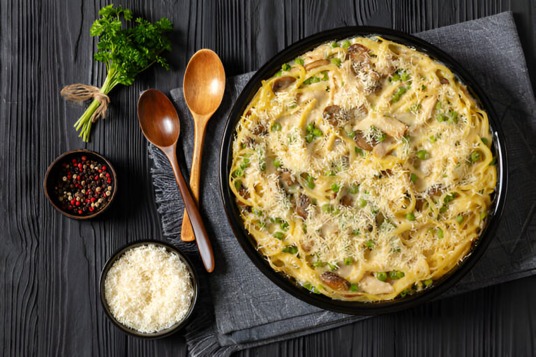 A bowl of creamy pasta topped with grated cheese, surrounded by parsley, wooden utensils, and spices.