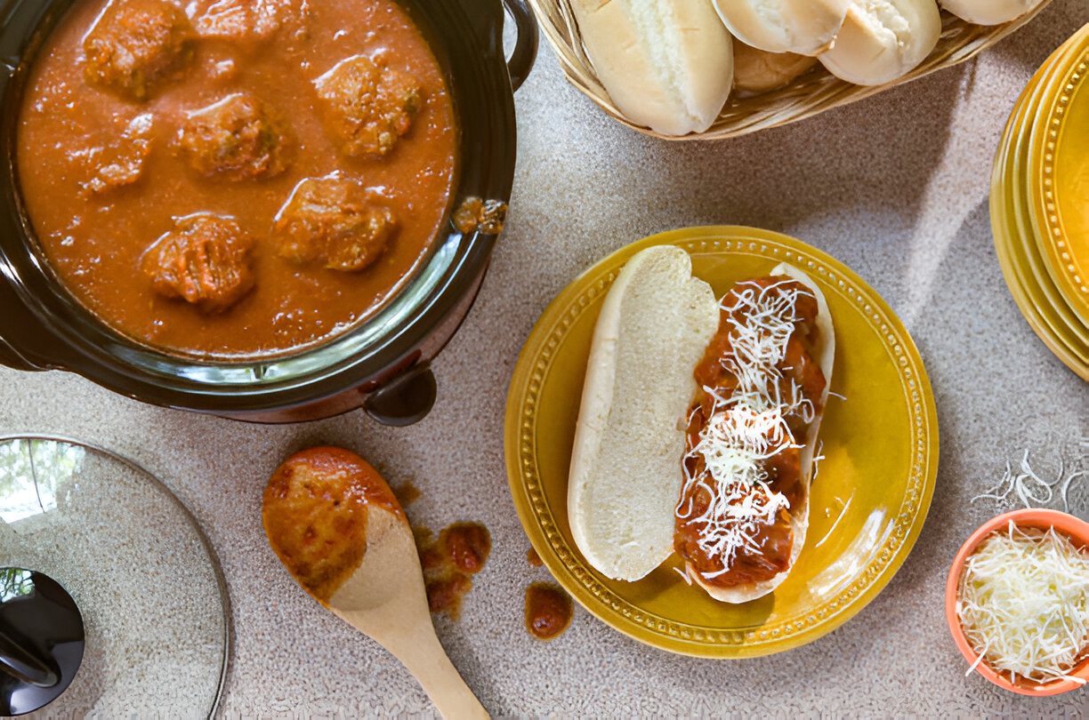 A plate of meatball sub with marinara sauce and shredded cheese, accompanied by a slow cooker filled with meatballs in sauce and a basket of buns.