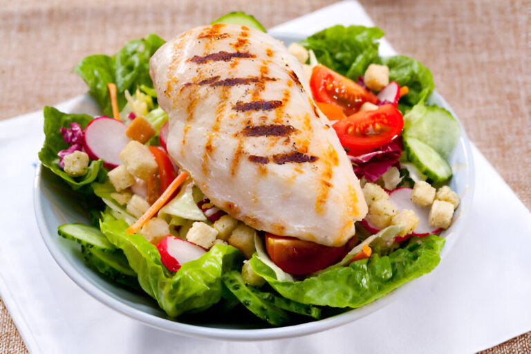 A delicious grilled chicken breast served on a bed of fresh salad with tomatoes, cucumbers, radishes, and croutons