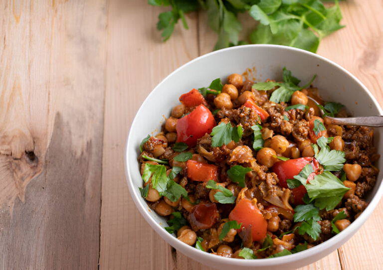 A bowl of chickpea and ground meat dish garnished with fresh parsley and diced tomatoes on a wooden table.