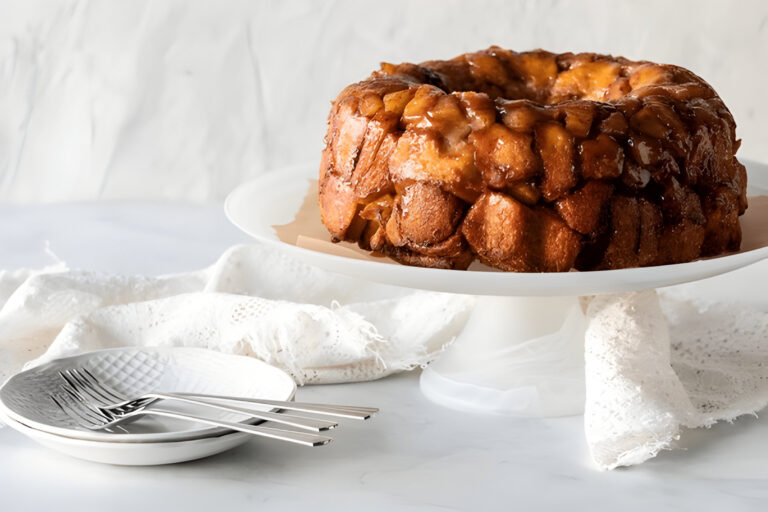 A golden, caramelized monkey bread on a white cake stand, surrounded by a white cloth and plates.