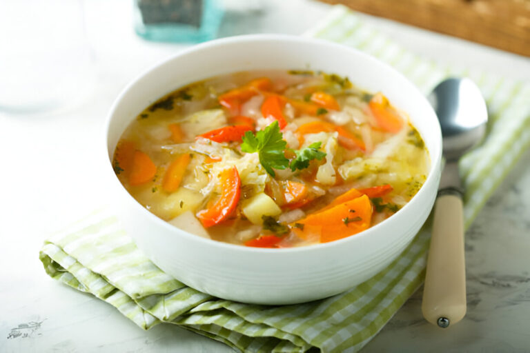 A bowl of colorful vegetable soup with carrots, red peppers, and cabbage on a green and white checkered napkin.