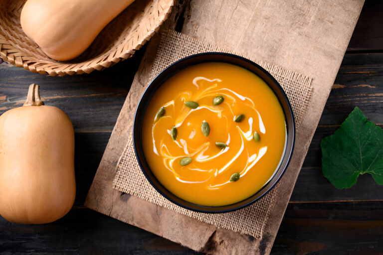 A bowl of creamy orange soup topped with pumpkin seeds, surrounded by butternut squash and leafy greens on a rustic wooden surface.