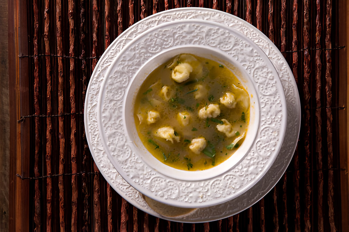 A bowl of soup with dumplings served on a decorative white plate over a textured brown mat.