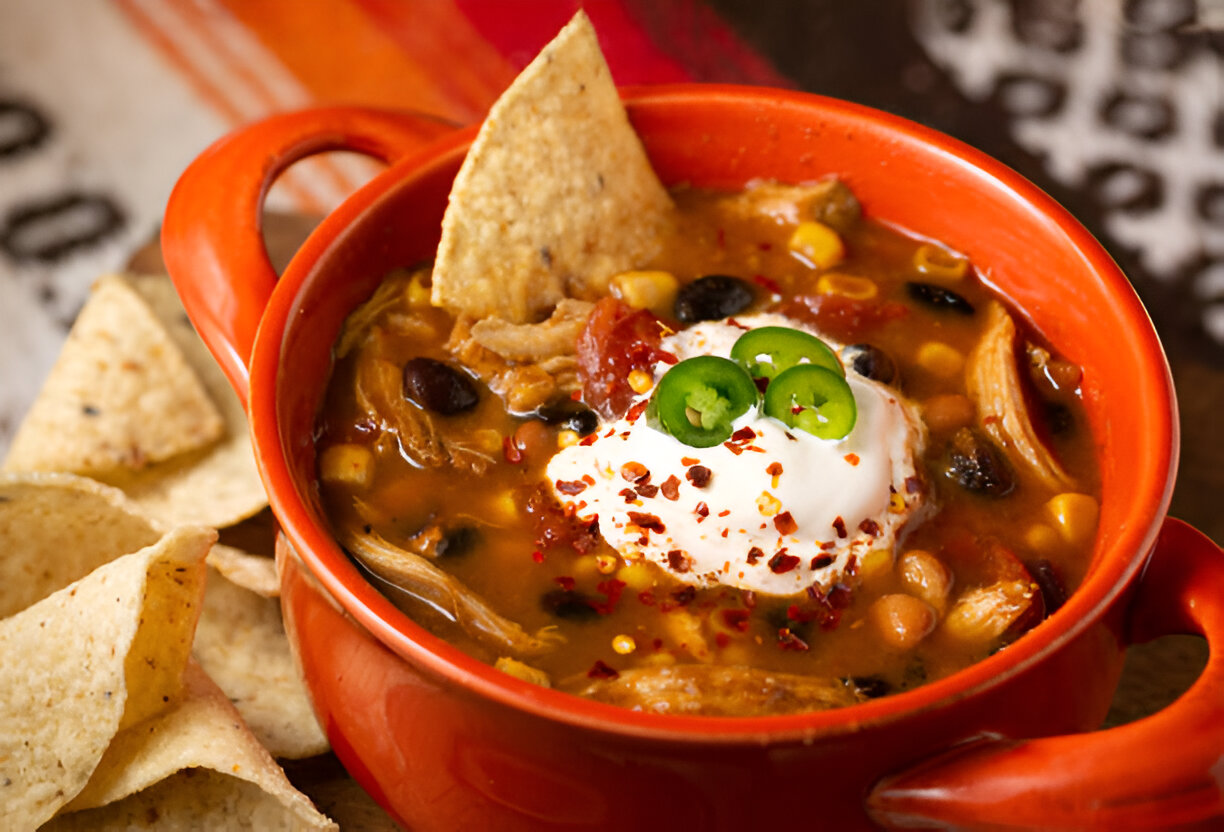 A vibrant orange bowl filled with a hearty chicken soup topped with sour cream, jalapeños, and spices, accompanied by tortilla chips.