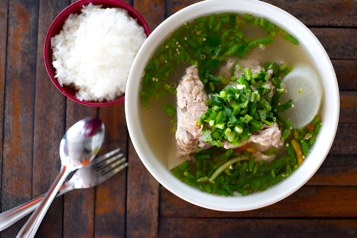 A bowl of soup with meat and green onions, accompanied by a side of white rice on a wooden table.