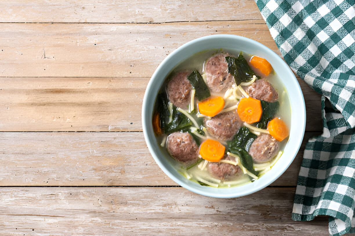 A bowl of meatball soup with carrots, greens, and noodles on a wooden table.