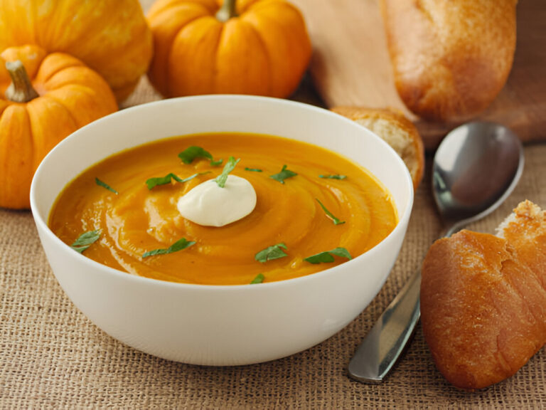 A bowl of creamy pumpkin soup topped with a dollop of sour cream and fresh herbs, accompanied by a piece of bread and small pumpkins.