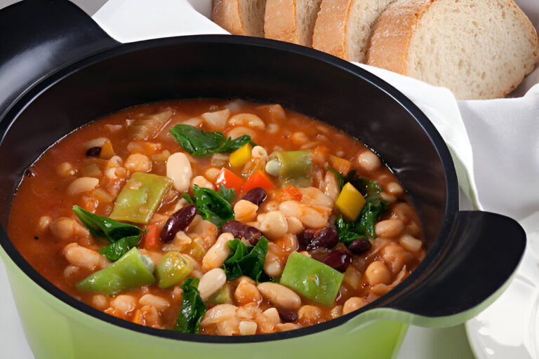 A colorful bean and vegetable stew served in a green pot with slices of bread in the background.