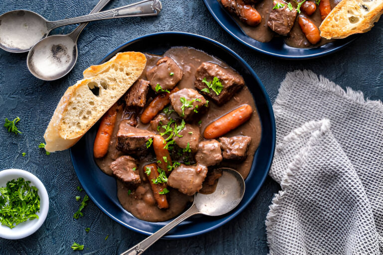 A close-up view of a hearty beef stew with carrots and herbs, served in a deep blue bowl alongside crusty bread and silver cutlery.