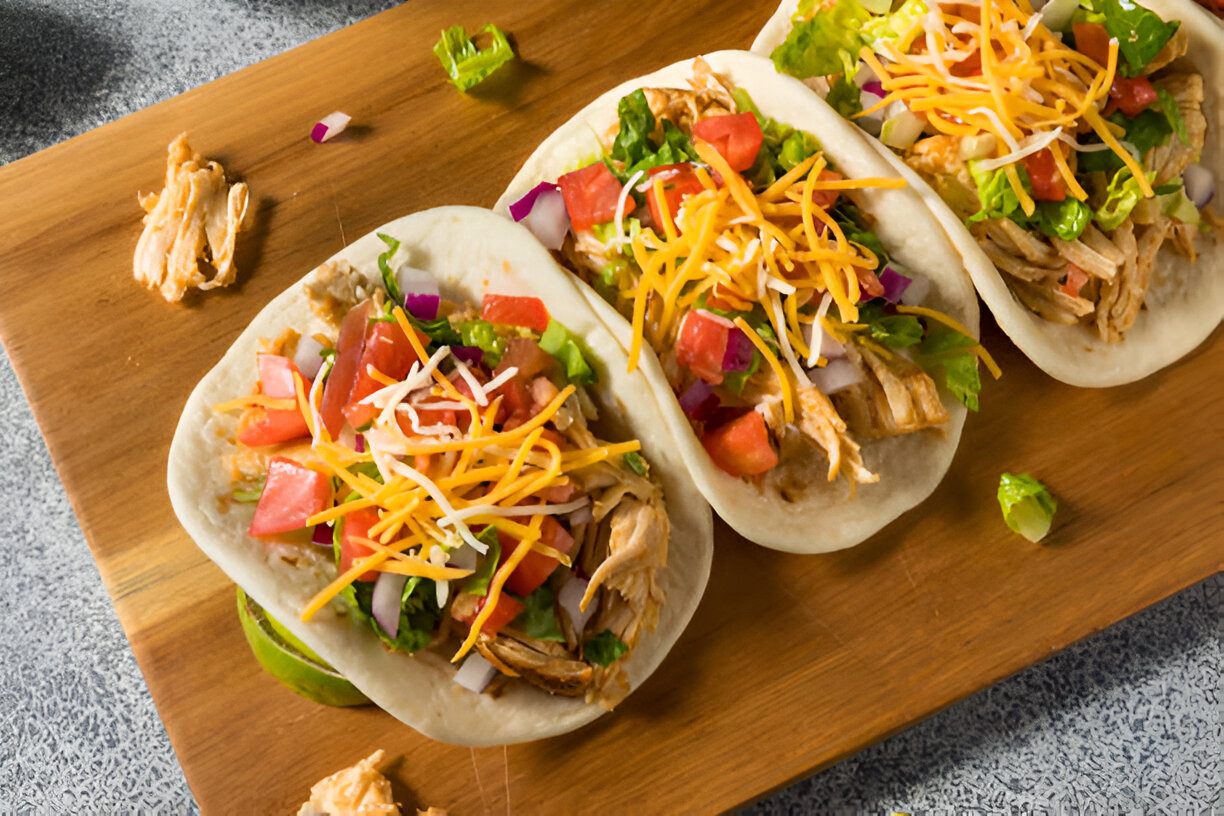 Three soft tacos filled with shredded chicken, lettuce, tomatoes, and cheese on a wooden cutting board.