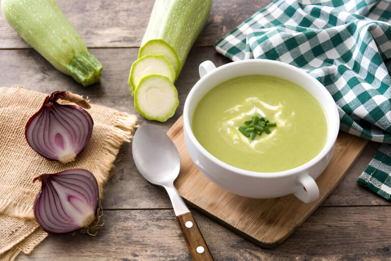 A bowl of green soup garnished with herbs, surrounded by chopped zucchini, red onions, and a green checkered napkin.