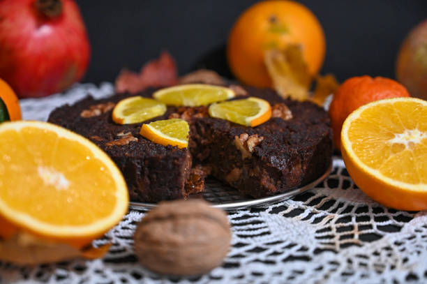 A delicious fruitcake garnished with orange slices, surrounded by fresh oranges and pomegranates on a lace tablecloth.