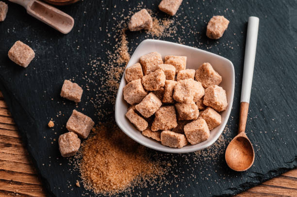 A bowl of brown sugar cubes on a dark slate surface with spilled granulated sugar and a wooden spoon.