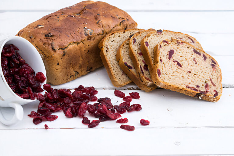 A loaf of cranberry bread with slices laid out next to scattered dried cranberries on a white wooden surface.