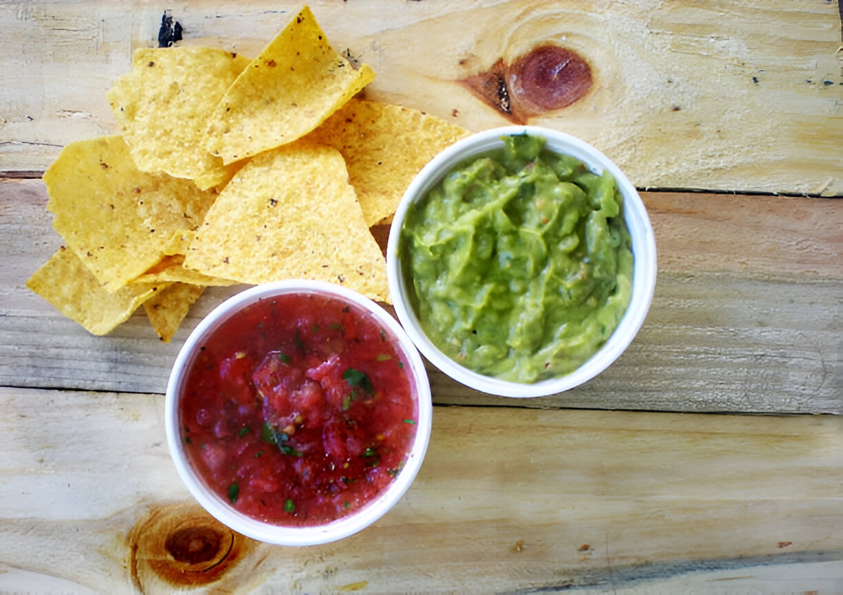 A plate of tortilla chips alongside two bowls of salsa and guacamole on a wooden surface.