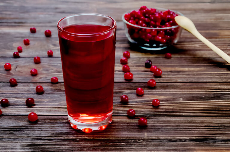 A glass of bright red drink on a wooden table, surrounded by fresh cranberries and a spoon.