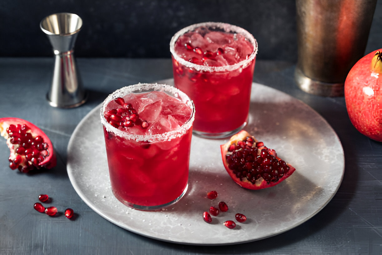 Two cocktails with pomegranate seeds on a serving tray, accompanied by fresh pomegranate slices and an alcohol measuring tool.