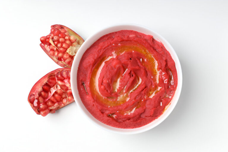 A bowl of red hummus swirled with olive oil, accompanied by a halved pomegranate.