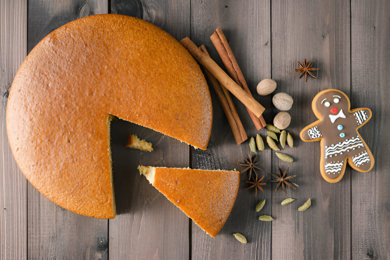 A round cake partially sliced, surrounded by spices and a gingerbread cookie on a wooden surface.