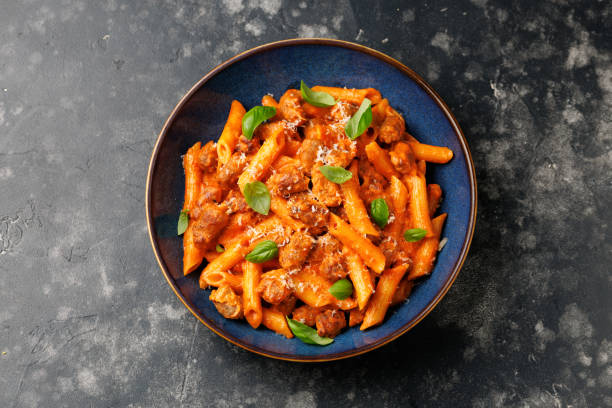 A bowl of penne pasta with tomato sauce, meatballs, and garnished with fresh basil leaves.