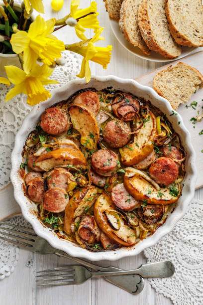 A delicious baked dish featuring sausage, potatoes, and onions, garnished with chopped herbs, served alongside slices of whole grain bread and yellow flowers.