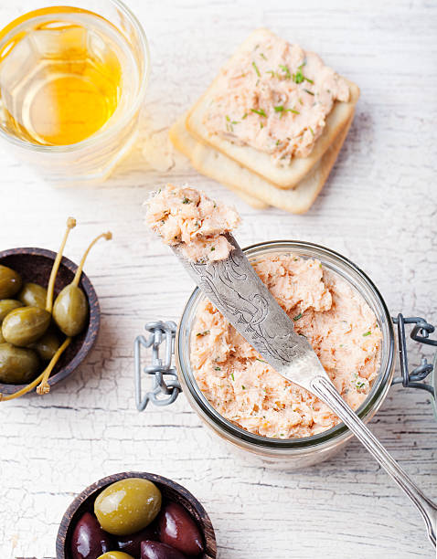 A jar of salmon pâté with a spreader, surrounded by olives, capers, crackers, and a glass of drink on a wooden table.