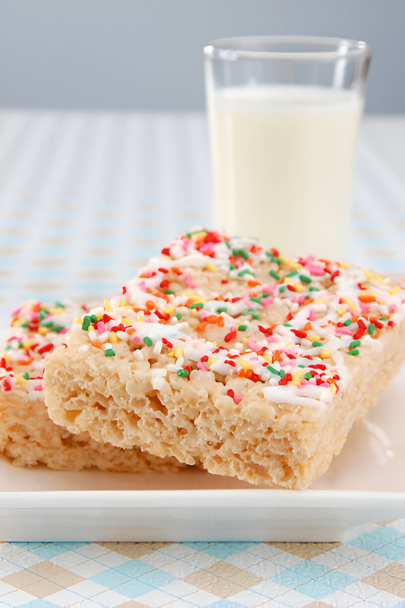 Two squares of colorful rice crispy treats topped with sprinkles on a plate, with a glass of milk in the background.