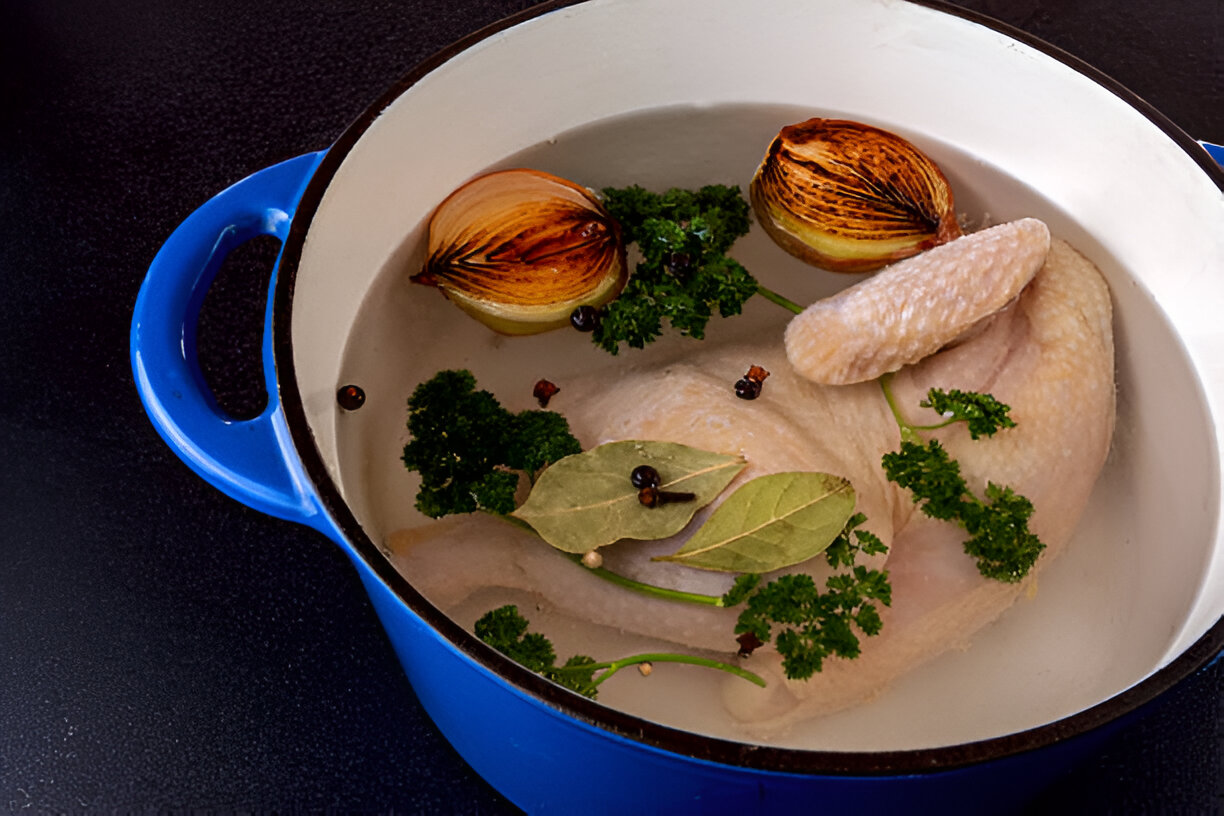 A fresh chicken submerged in water with herbs and spices in a blue pot.