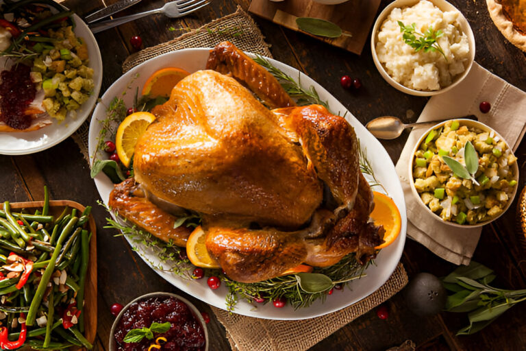 A beautifully roasted turkey garnished with orange slices and fresh herbs, surrounded by various sides including green beans, stuffing, and mashed potatoes.