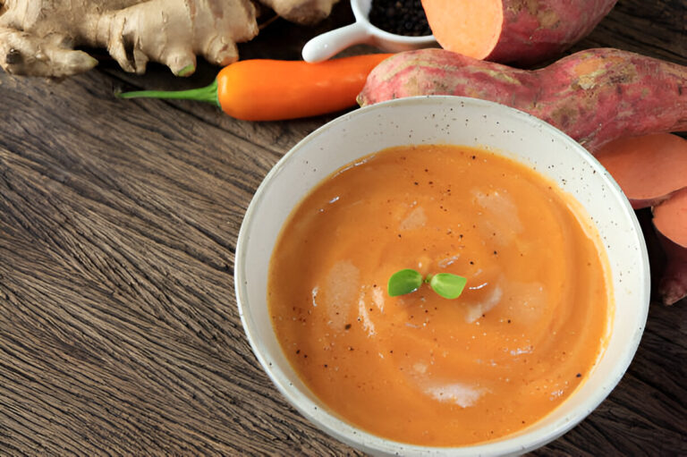 A creamy orange soup garnished with herbs, surrounded by fresh ginger and sweet potatoes on a wooden table.