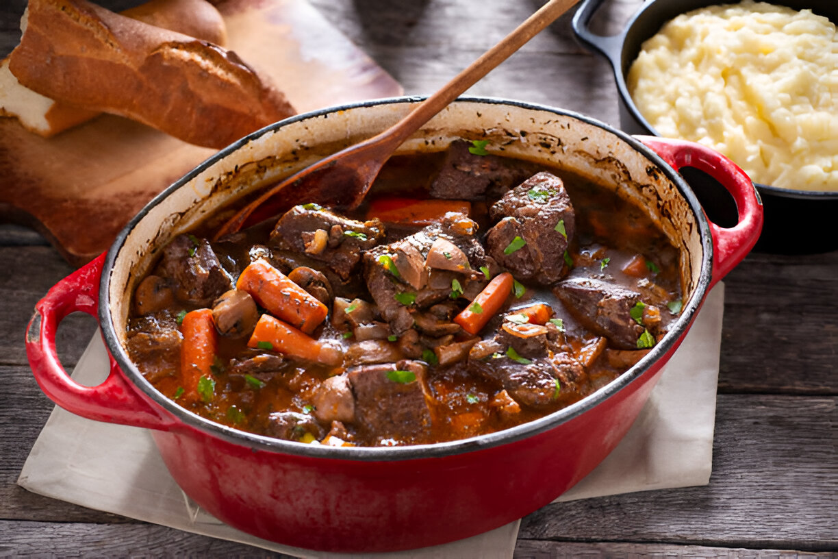 A delicious beef stew with carrots and mushrooms in a red pot, accompanied by a side of mashed potatoes and crusty bread.