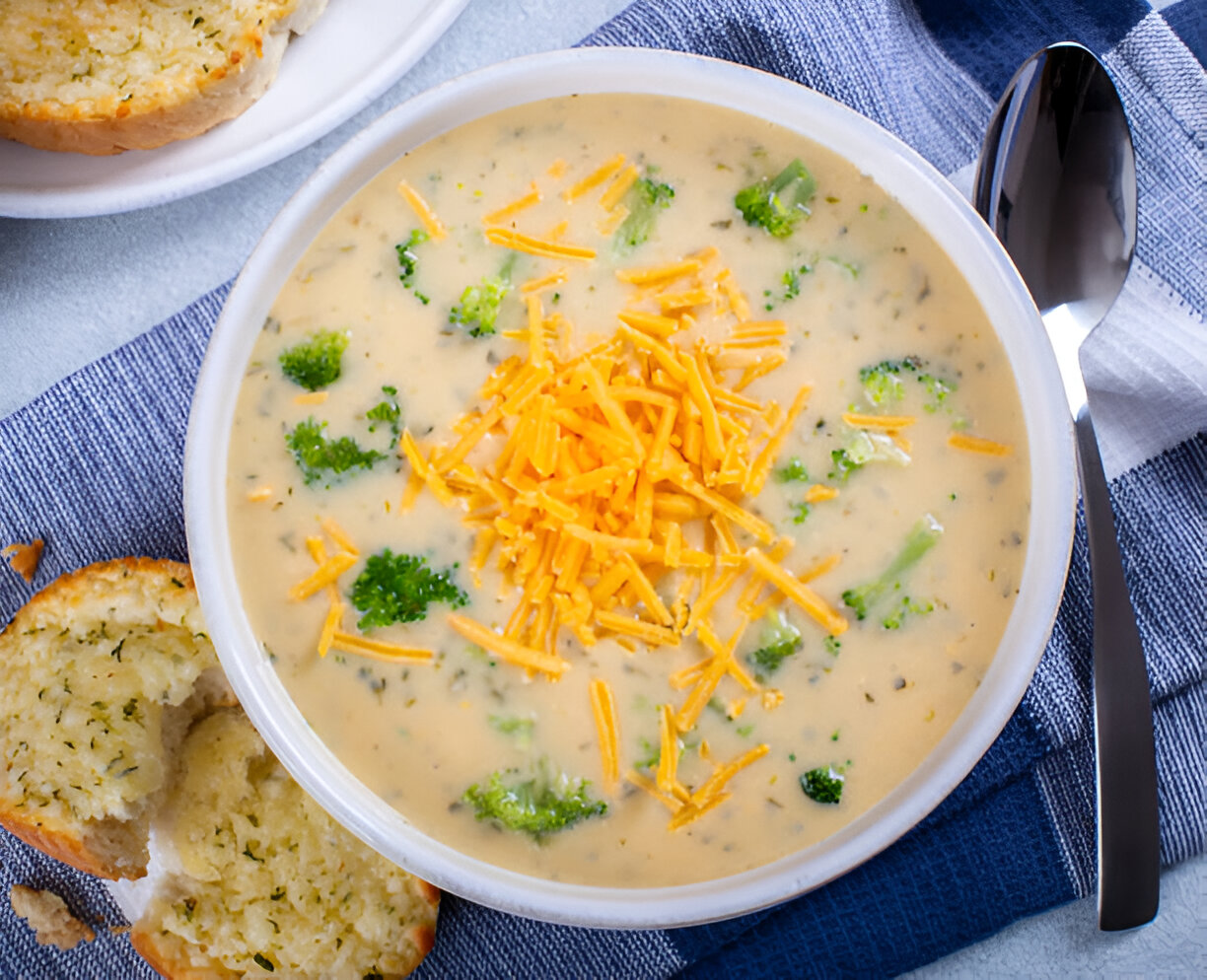 Creamy broccoli cheese soup in a white bowl topped with shredded cheddar cheese, served with pieces of bread on the side.