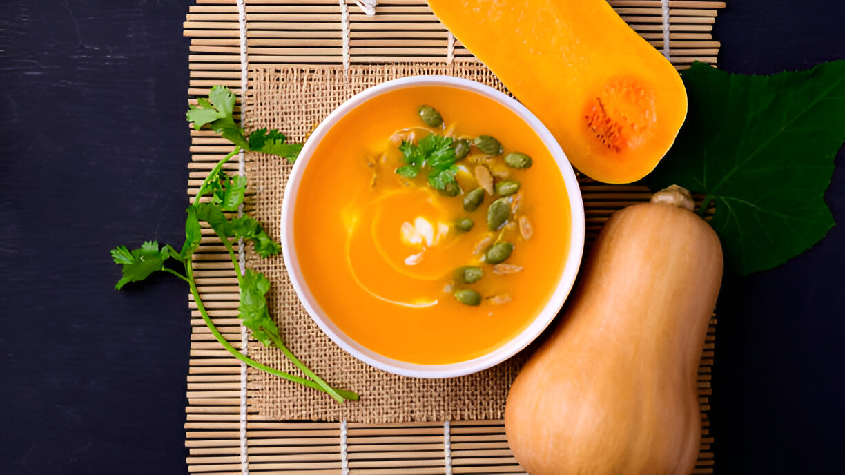 A bowl of vibrant orange butternut squash soup garnished with pumpkin seeds and cilantro, accompanied by fresh butternut squash.