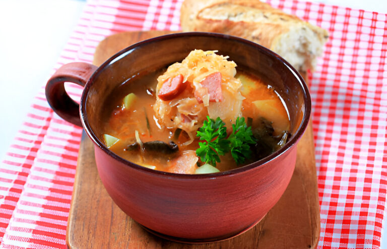 A bowl of hearty soup filled with vegetables and sausage, garnished with parsley, on a wooden cutting board with a piece of bread in the background.