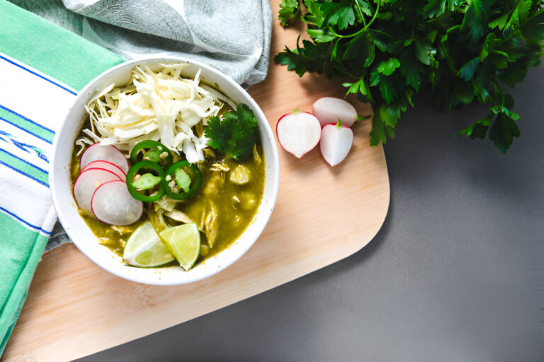 A bowl of green soup topped with cabbage, radishes, jalapeños, and cilantro, alongside fresh lime and radishes on a wooden cutting board.