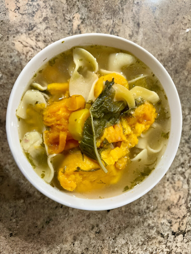 A bowl of hearty soup with vegetables and noodles on a textured countertop.