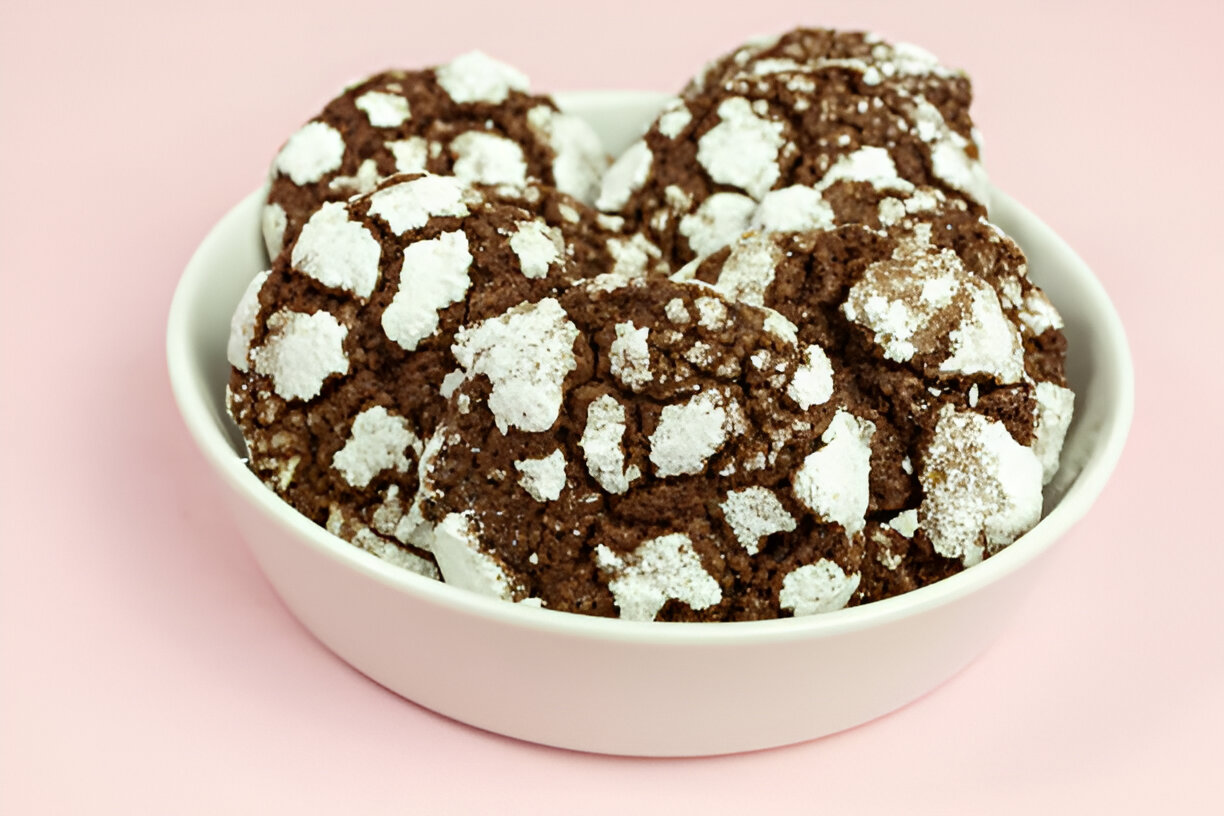 A bowl of chocolate crinkle cookies with powdered sugar on a pink background.