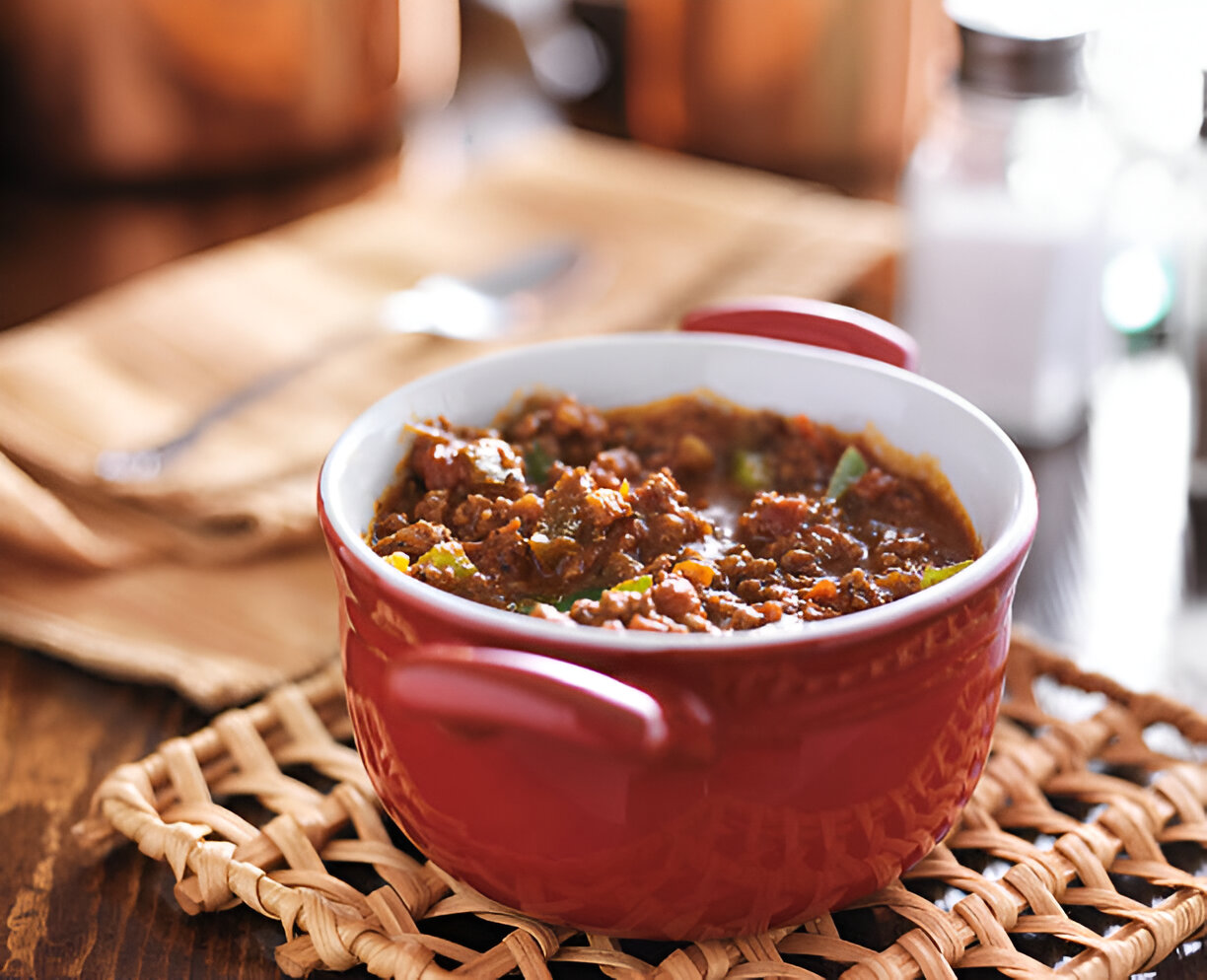 A red bowl of chili with green peppers on a woven mat