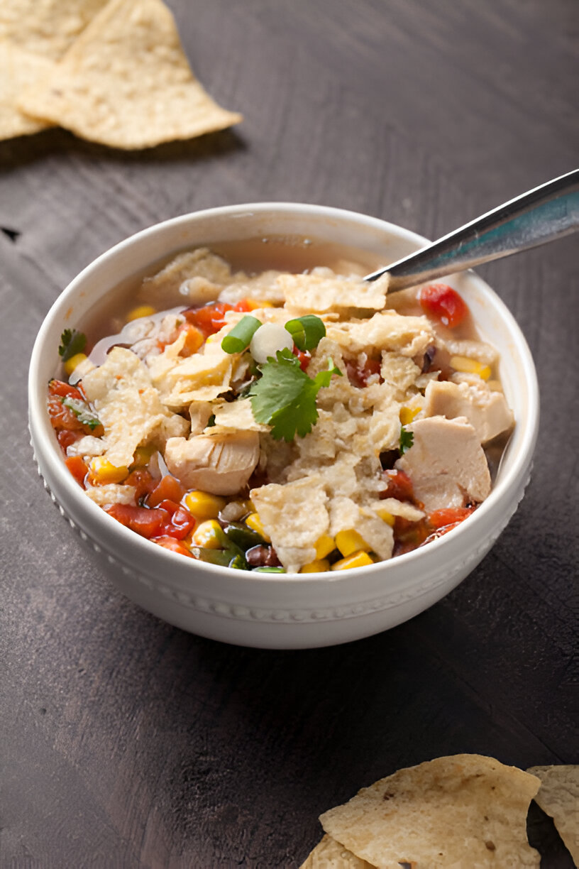 A bowl of chicken soup topped with crushed tortilla chips, diced tomatoes, corn, beans, and garnished with cilantro and green onions.