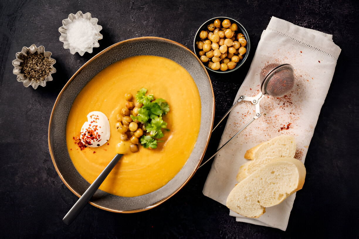 A bowl of creamy orange soup topped with chickpeas and fresh cilantro, served with a dollop of sour cream and a side of slices of bread.