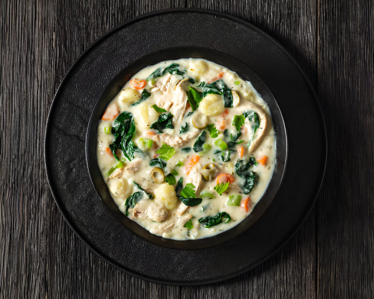 A bowl of creamy chicken and vegetable soup with spinach, carrots, and herbs on a dark wooden surface.