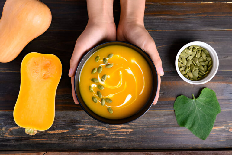 A bowl of creamy butternut squash soup held in hands, surrounded by a halved butternut squash, pumpkin seeds, and a green leaf, on a wooden table.