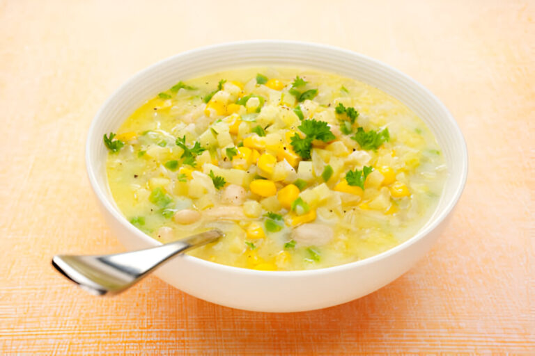 A bowl of creamy corn soup garnished with cilantro and green onions, served with a spoon on an orange textured surface.