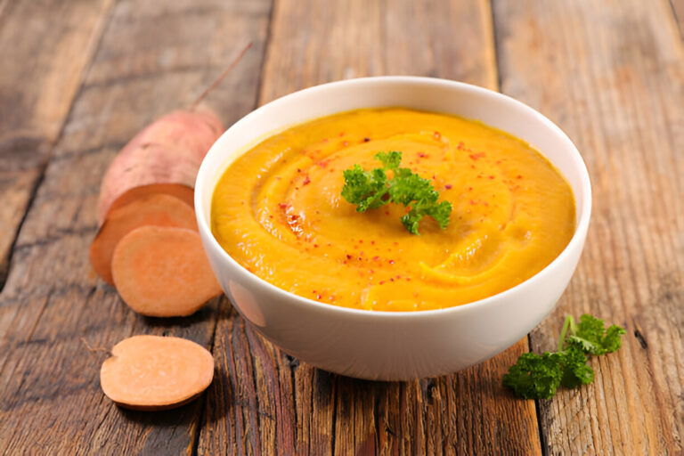 A bowl of creamy sweet potato soup garnished with parsley on a wooden table, with sliced sweet potatoes nearby.