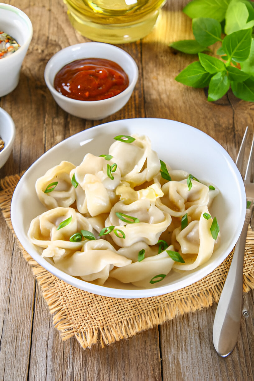 A white bowl filled with dumplings topped with green onions, surrounded by small bowls of sauces on a rustic wooden table.